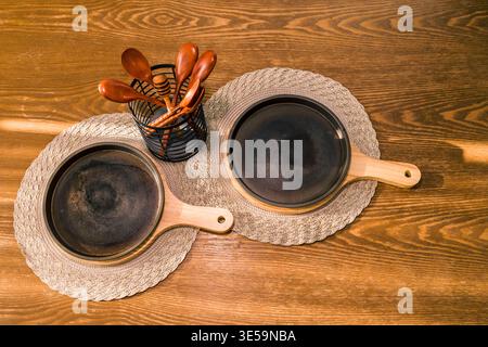 Symmetrical arrangement of kitchenware includes two small cast-iron skillets resting on grey woven placemats on a wooden table. Stock Photo