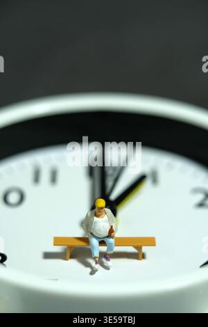 Miniature people toy figure photography. Fat men worker take a break rest above clock. Image photo Stock Photo