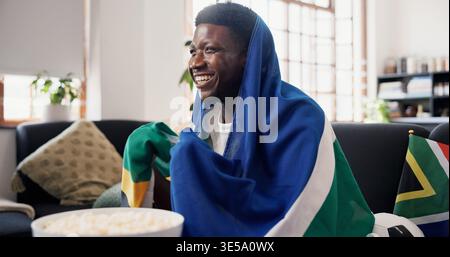 Man, watching tv and South Africa flag for soccer on couch with goals, excited and support team in home. African person, smile and snack on sofa Stock Photo