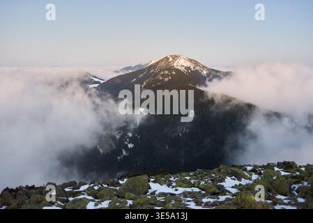 Breathtaking photograph showcases majestic mountain peak, partially covered with snow and surrounded by dense pine forests. Mist and clouds weave through valleys under clear blue sky. Stock Photo