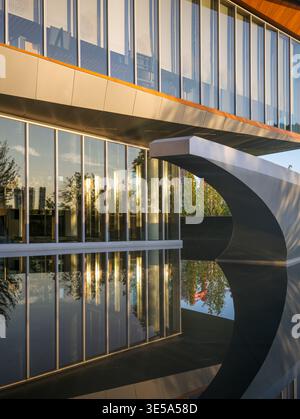 A modern commercial building featuring expansive glass windows, a sleek curved architectural overhang, and wooden ceiling details, flawlessly reflecte Stock Photo