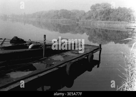 Sloviansk, November 7, 1996, Sloviansk Resort. A captivating close-up view of weathered metal pontoon boats quietly moored at the muddy shore of a calm resort lake. The still water perfectly mirrors the distant dense tree line wrapped in a delicate autumn haze, while tall wild reeds border the right side of the frame. This highly evocative and moody black and white archival picture elegantly documents the quiet, untouched nature of the area, beautifully capturing life before the war in a completely peaceful Donbas. Stock Photo