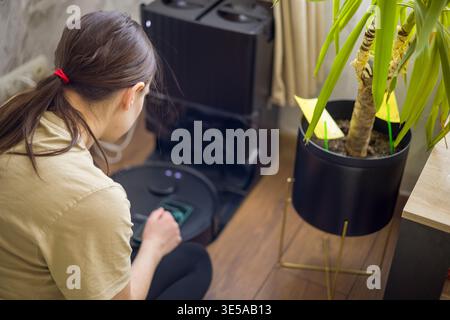 Woman Using App To Control Robot. Woman Operates Robotic Cleaner Via Application Interface. Female User Adjusts Device Settings With Smartphone Near Stock Photo