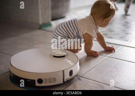 Infant Explores Modern Home Robot With Cautious Fascination. Caucasian Baby Carefully Inspects Robotic Vacuum Cleaner On Tiled Surface With Playful Stock Photo