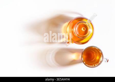 Top view of glass cup of tea and teapot with long shadows on white background. Stock Photo