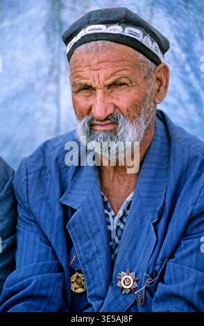 An elderly man sits in Bukhara, Uzbekistan. He wears traditional clothes and appears to be engaging with others nearby. Stock Photo