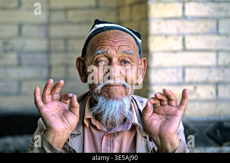 An elderly man sits in Khiva, Uzbekistan and displays hand gestures with his hands raised. He smiles warmly at passersby. Stock Photo