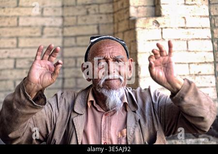 An elderly man raises his hands while interacting with visitors in Khiva, Uzbekistan. The sun shines on the brick walls around him. Stock Photo
