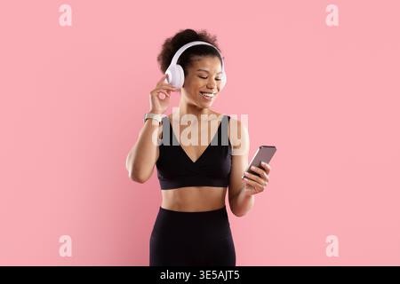 Smiling black sportswoman in activewear adjusting white headphones while looking at smartphone Stock Photo