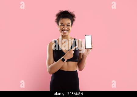 Smiling black sportswoman in activewear pointing at smartphone with blank white screen Stock Photo