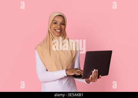 Young black Muslim woman in hijab smiling confidently while holding open laptop Stock Photo
