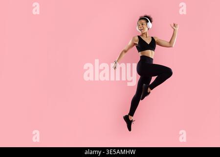 Happy black woman jumping with raised arms and headphones on pink background Stock Photo