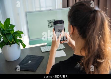 Person scanning QR code from computer screen with smartphone at home workspace. Digital interaction between devices and online service access concept. Stock Photo