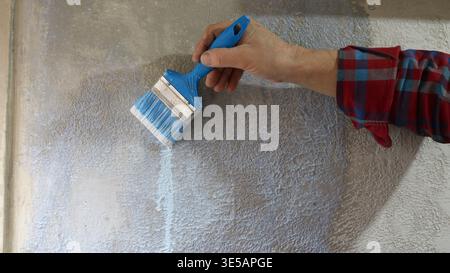Close up of man in plaid shirt holding blue paintbrush applying liquid primer to textured grey plaster wall and priming wall of house Stock Photo