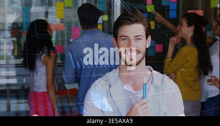 Smiling man in hoodie holding teal marker at office meeting, glass wall notes and digital charts Stock Photo