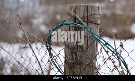 A weathered wooden post holds up a rusted chain-link fence with a loop of blue wire against a blurred winter background. Stock Photo