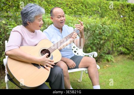 Senior woman sitting on a bench in the garden playing an acoustic guitar while a friend teaching. Concept of music learning and creative leisure in retirement Stock Photo