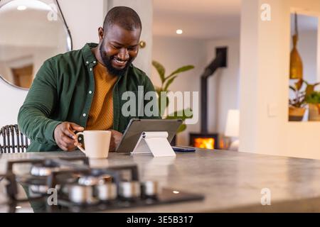 African American man smiling, using tablet on white adjustable stand at kitchen island, holding mug. Device, adult, homeoffice, remote, modern, interiors, openplan Stock Photo