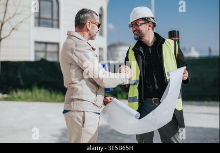 Construction engineer and contractor shaking hands on site while reviewing blueprints and plans Stock Photo