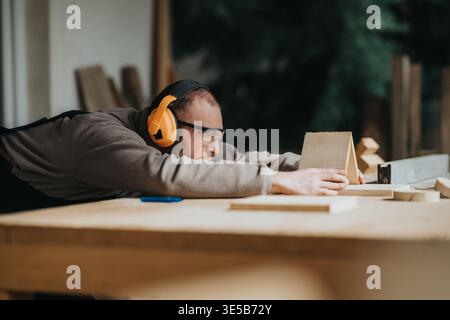 Carpenter inspecting and measuring a wooden piece wearing safety glasses and ear protection in workshop Stock Photo
