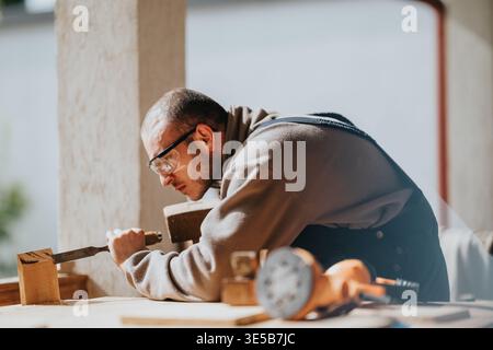 Carpenter carefully carving wood with a chisel in a bright workshop, concentrating on detail Stock Photo