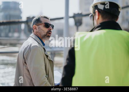 Construction manager discussing plans with site worker wearing safety vest and hard hat by waterfront Stock Photo