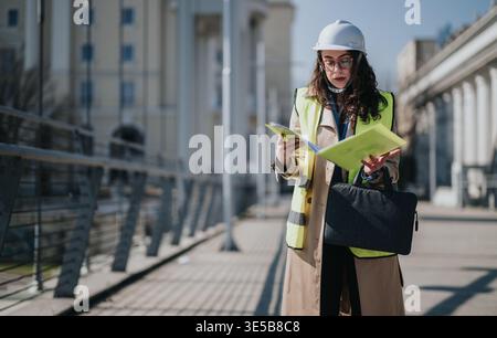 Female engineer inspecting urban construction site in hard hat and high-visibility vest with plans Stock Photo