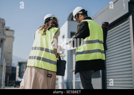 Two construction engineers in safety vests and hard hats reviewing building plans at a site Stock Photo