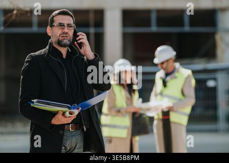 Construction engineers on site with manager speaking on phone and colleagues reviewing plans Stock Photo