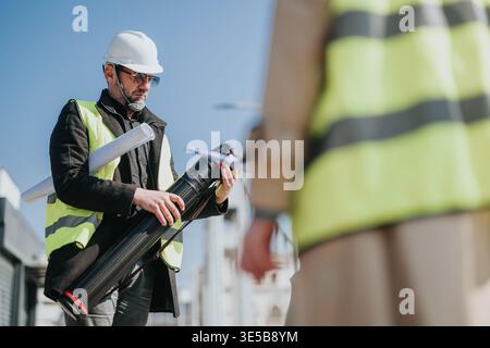 Construction engineer in hard hat and safety vest holding blueprints and plan tube on site Stock Photo