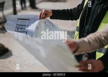 Architects reviewing construction blueprints on site while wearing high-visibility vests and discussing plans Stock Photo