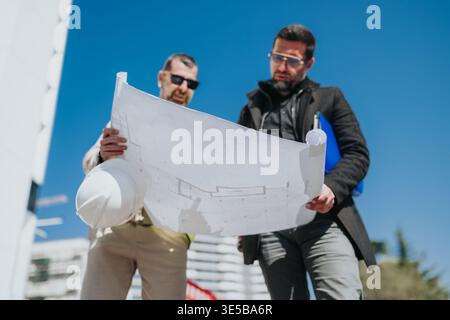Two architects reviewing blueprints on a construction site during a sunny urban planning meeting Stock Photo