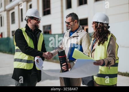 Construction professionals shaking hands over blueprints at building site wearing hard hats and safety vests Stock Photo