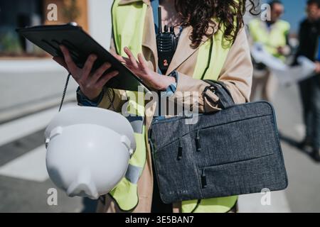 Female construction manager in safety vest using tablet with hard hat and briefcase on site Stock Photo