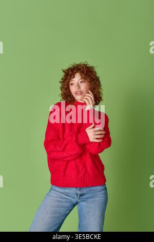 A young woman with stunning red curls poses gracefully in a cozy red sweater on green. Stock Photo