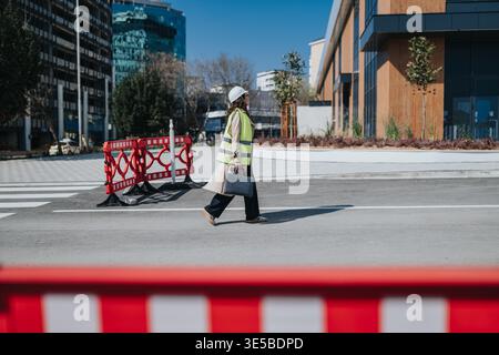 Female construction engineer in safety vest and hard hat walking across urban street with briefcase Stock Photo