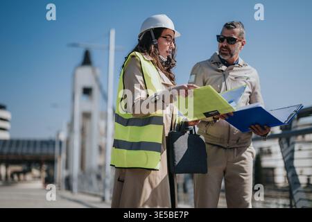 Female and male engineers discussing construction plans on site wearing hardhat and high-visibility vest Stock Photo