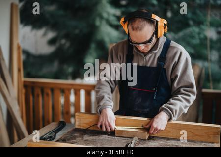 Carpenter measuring a wooden plank at a workshop table wearing safety gear Stock Photo