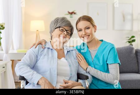 Portrait of young cheerful nurse with senior disabled patient in wheelchair looking at camera. Stock Photo