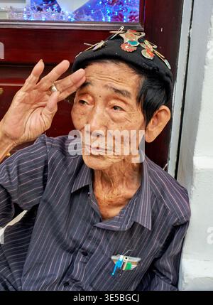 A man sits on the street in Ho Chi Minh City. He salutes with a hat decorated with pins. Street life continues around him. Stock Photo