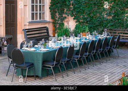 A long formal dining table with a green tablecloth set up outside on a brick patio with black chairs, wine glasses, and plates in front of a building Stock Photo