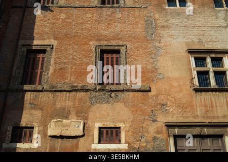 Historic building facade in Rome showing aged stucco, brown shutters, stone window frames, and an inscribed marble plaque Stock Photo
