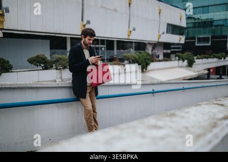 Businessman standing in urban plaza checking smartphone while holding a red bag and leaning on railing Stock Photo