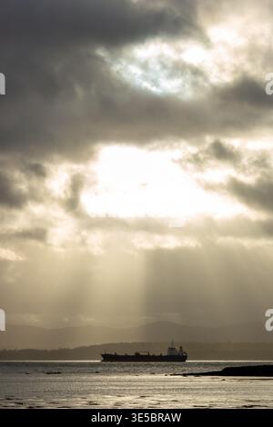 A cargo ship in the distance on the Firth of Forth under a dramatic, cloudy sky with sunbeams breaking through Stock Photo