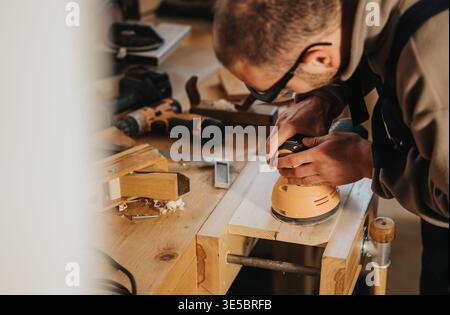 Carpenter sanding wooden plank in workshop using electric sander on workbench Stock Photo
