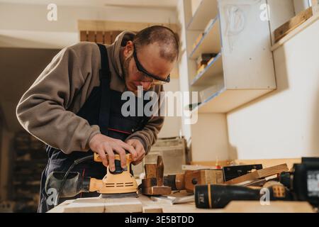 Carpenter sanding wood on workbench in workshop using electric sander and protective glasses Stock Photo