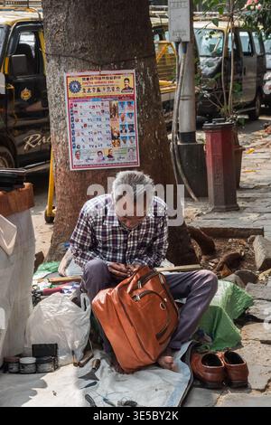 A cobbler sitting by the roadside pavement. Shoemaker repairs shoes and bags right on the street of Mumbai India. Senior man repairing a leather bag o Stock Photo