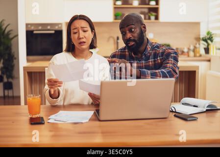A couple sits at a table reviewing bills and documents, looking concerned as they compare paperwork with information on a laptop. Stock Photo