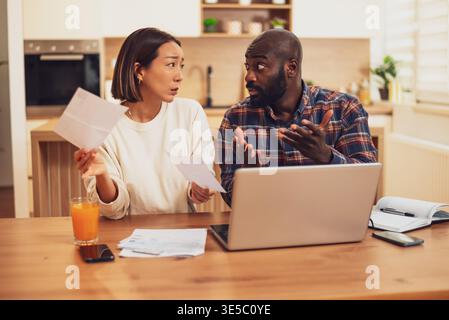 A couple sits at a table reviewing bills and documents, looking concerned as they compare paperwork with information on a laptop. Stock Photo