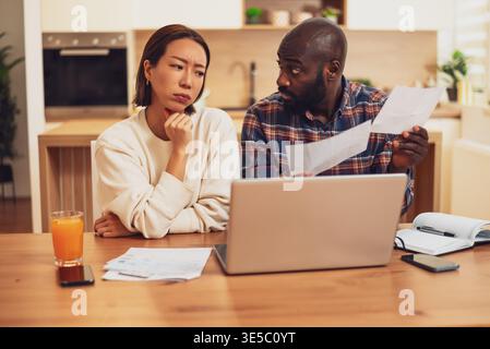 A couple sits at a table reviewing bills and documents, looking concerned as they compare paperwork with information on a laptop. Stock Photo